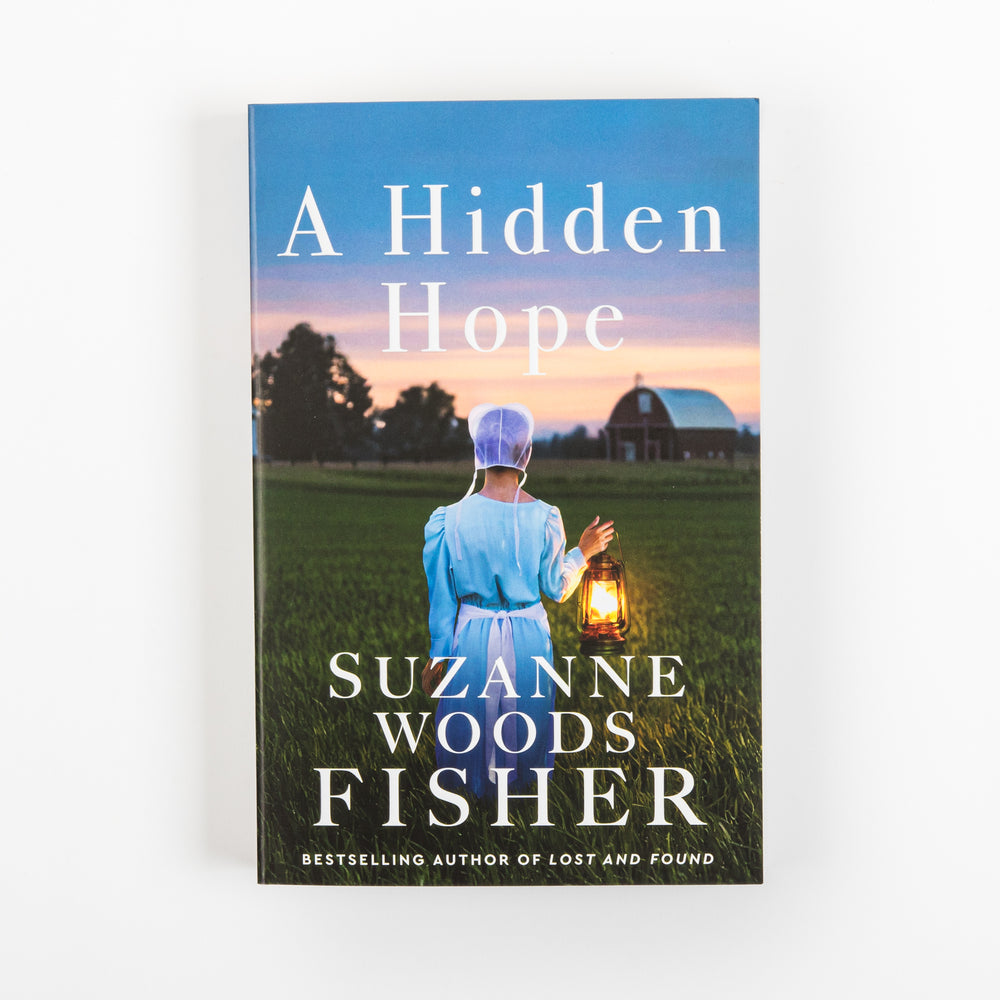 Front cover of A Hidden Hope by Suzanne Woods Fisher, featuring an Amish woman in a bonnet and blue dress holding a glowing lantern in a green field at sunset, with a red barn behind her; cover notes she is the bestselling author of Lost and Found.