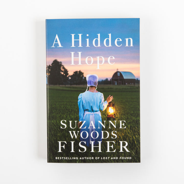 Front cover of A Hidden Hope by Suzanne Woods Fisher, featuring an Amish woman in a bonnet and blue dress holding a glowing lantern in a green field at sunset, with a red barn behind her; cover notes she is the bestselling author of Lost and Found.