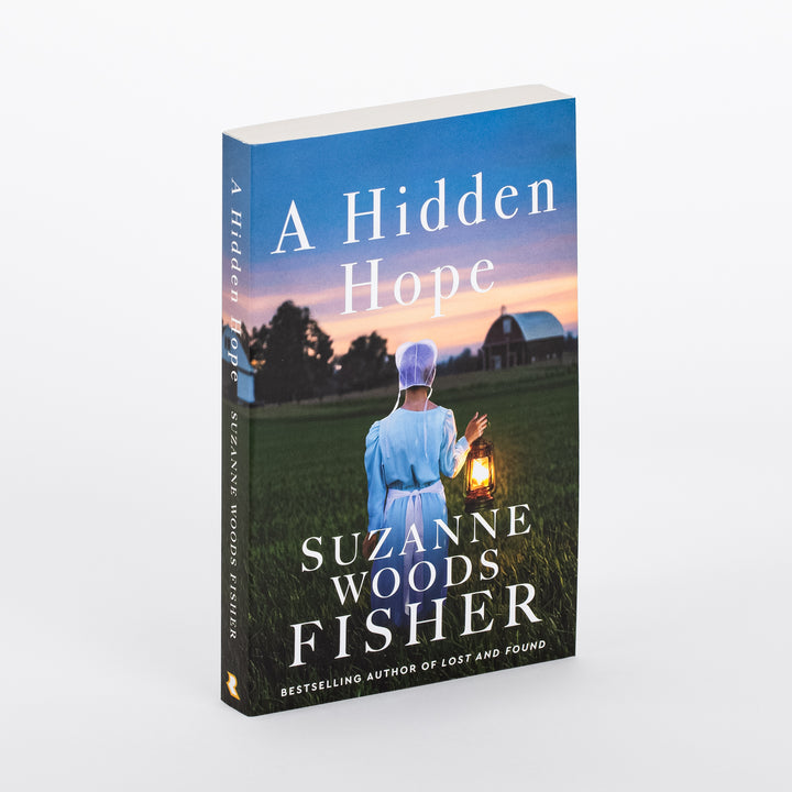 Angled 3/4 view of the paperback cover of A Hidden Hope by Suzanne Woods Fisher, showing an Amish woman in a bonnet and blue dress carrying a lit lantern through a green field at dusk, with a red barn in the background.