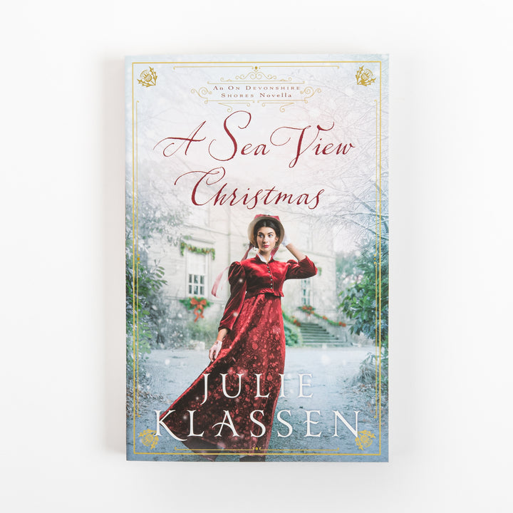 Front cover of the paperback novel "A Sea View Christmas" by Julie Klassen, featuring a woman in a red velvet gown and bonnet walking past a snowy stone manor house trimmed with holiday greenery, framed by a gold Victorian-style border.