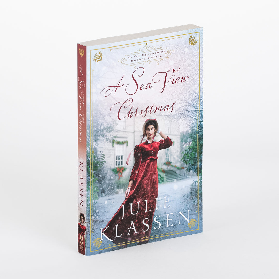 Angled paperback cover of "A Sea View Christmas" by Julie Klassen, showing a young woman in a red Victorian-style dress and bonnet standing before a snow-dusted English manor decorated with Christmas wreaths and garlands.