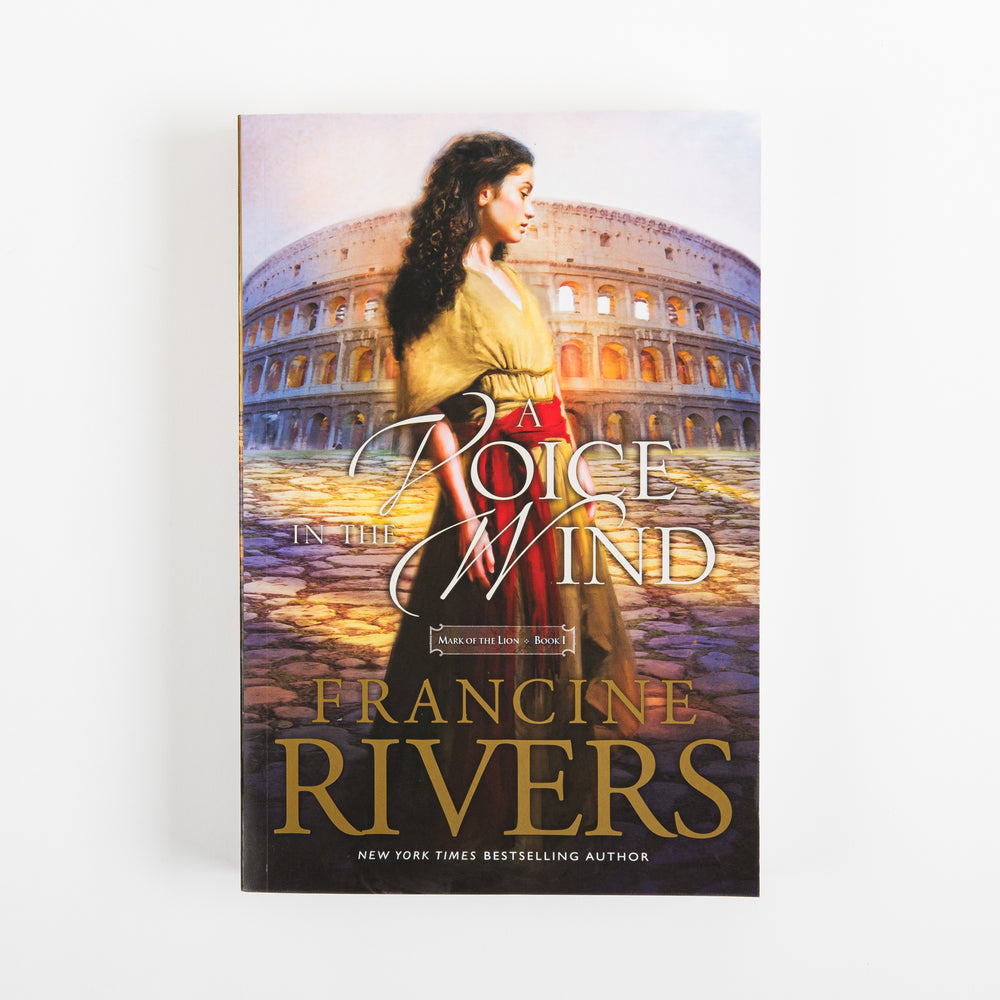Front cover of the paperback A Voice in the Wind by Francine Rivers, Mark of the Lion series Book 1, shown flat and straight-on. A young woman with long dark curly hair, in a gold blouse and red sash, stands with her back turned in front of the glowing ancient Roman Colosseum at twilight. The title appears in white cursive script with the series and book label beneath it, and the author's name in bold gold capital letters below, followed by a New York Times bestselling author line.