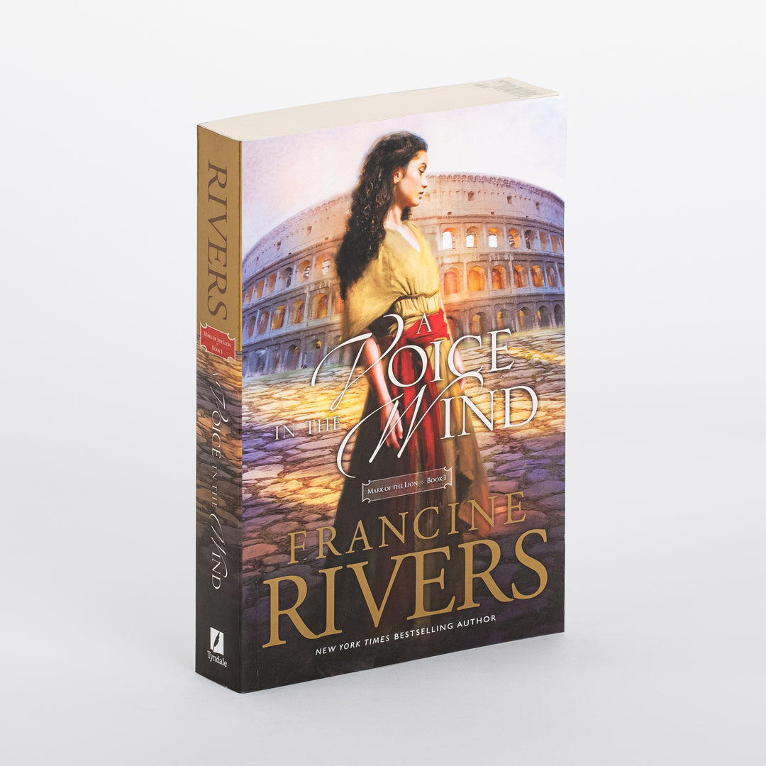 Paperback of A Voice in the Wind by Francine Rivers, shown at a three-quarter angle so both the front cover and spine are visible. The cover art shows a dark-haired woman in a gold blouse and red skirt standing before the ancient Roman Colosseum at dusk, with the title in white script and 'Mark of the Lion, Book 1' below it. The author's name appears in large gold letters with a New York Times bestselling author tagline; the spine reads RIVERS with a red ribbon marking the edition.