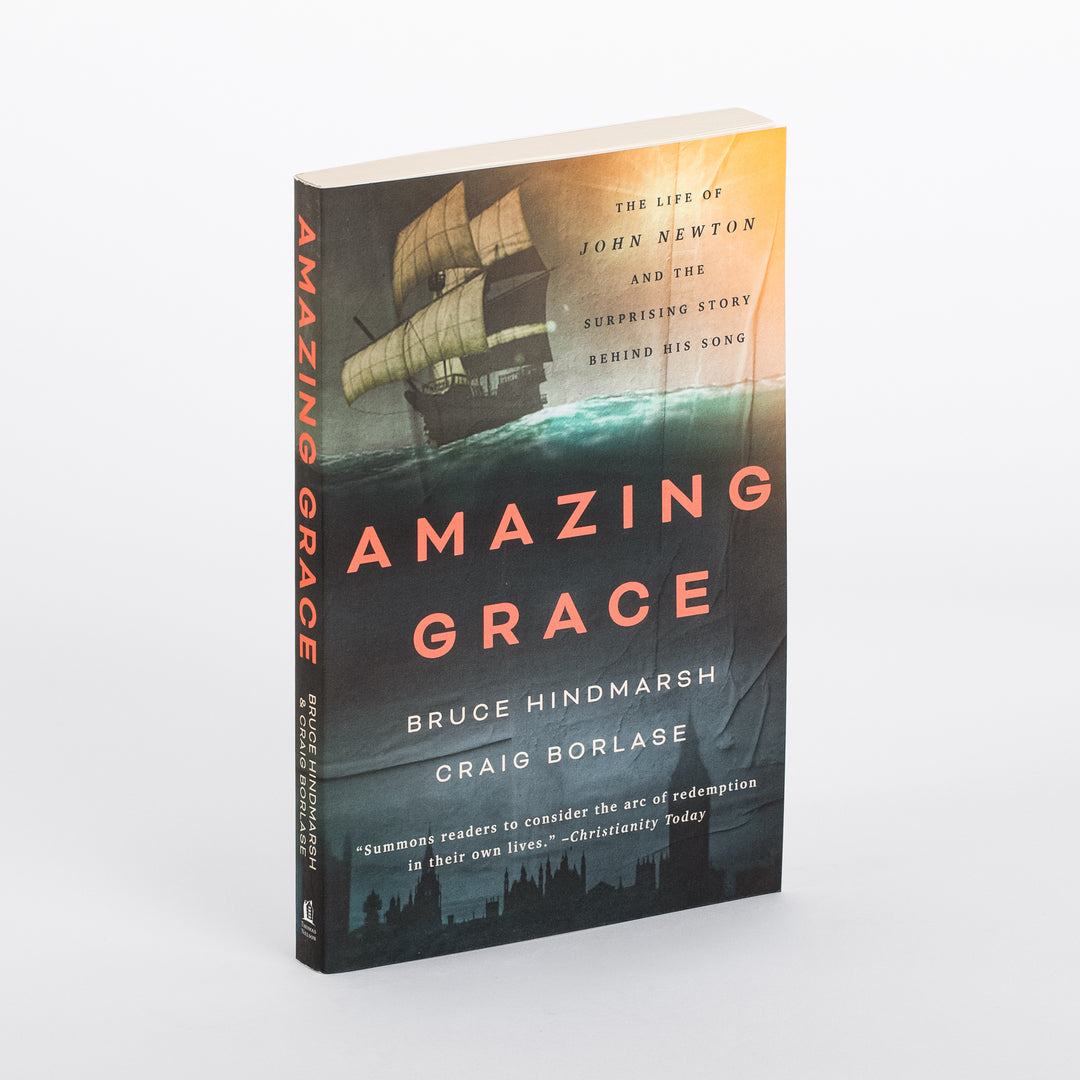 Paperback copy of Amazing Grace: The Life of John Newton and the Surprising Story Behind His Song standing upright at an angle, showing spine and front cover with a sailing ship illustration over teal ocean waves and an orange sunset sky.