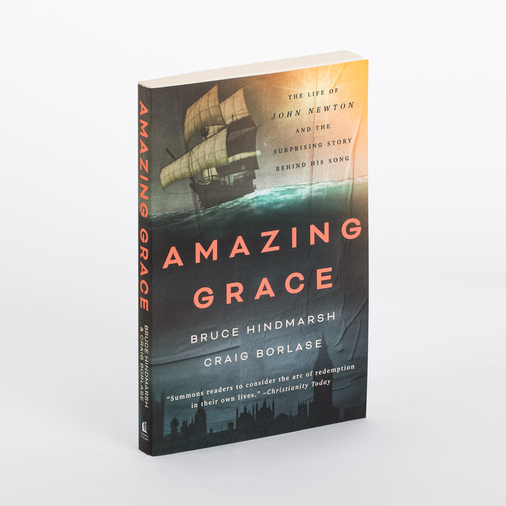 Paperback copy of Amazing Grace: The Life of John Newton and the Surprising Story Behind His Song standing upright at an angle, showing spine and front cover with a sailing ship illustration over teal ocean waves and an orange sunset sky.