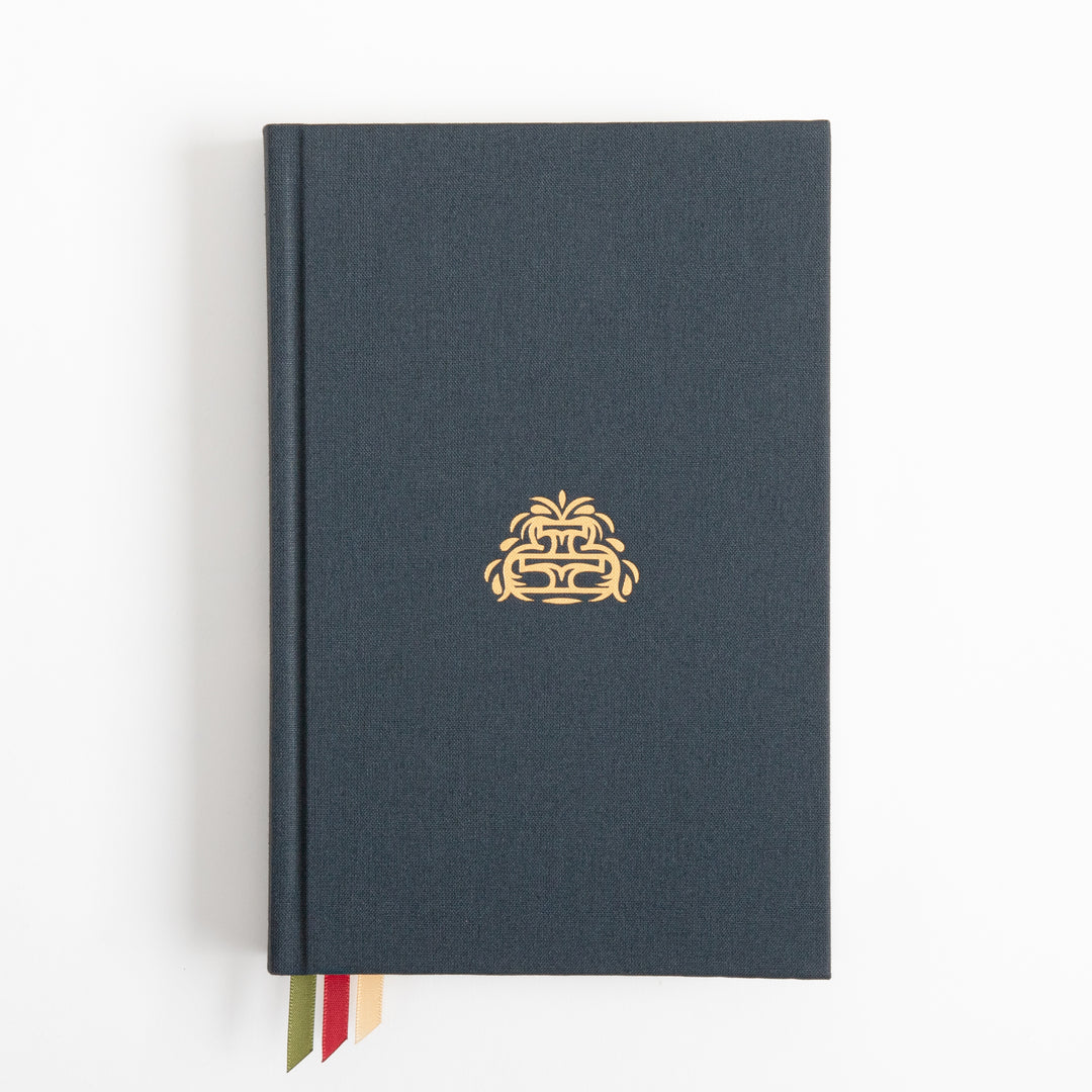 Overhead flat-lay of the closed hardcover book, bound in plain slate-blue cloth with a single small gold fountain emblem centered on the cover and no other text visible; three ribbon markers in cream, red, and olive green peek out from the bottom edge.