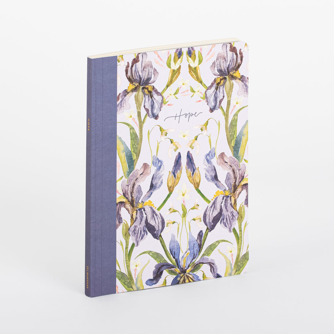 Angled view of the ESV Prayer Journal: 30 Days on Hope softcover journal, showing its blue linen spine and cream cover printed with a symmetrical, mirrored watercolor illustration of purple, lavender, and blue iris flowers with gold accents, and the word 'Hope' in cursive script centered at the top.