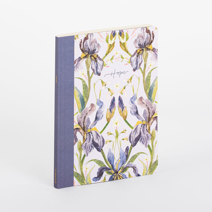 Angled view of the ESV Prayer Journal: 30 Days on Hope softcover journal, showing its blue linen spine and cream cover printed with a symmetrical, mirrored watercolor illustration of purple, lavender, and blue iris flowers with gold accents, and the word &