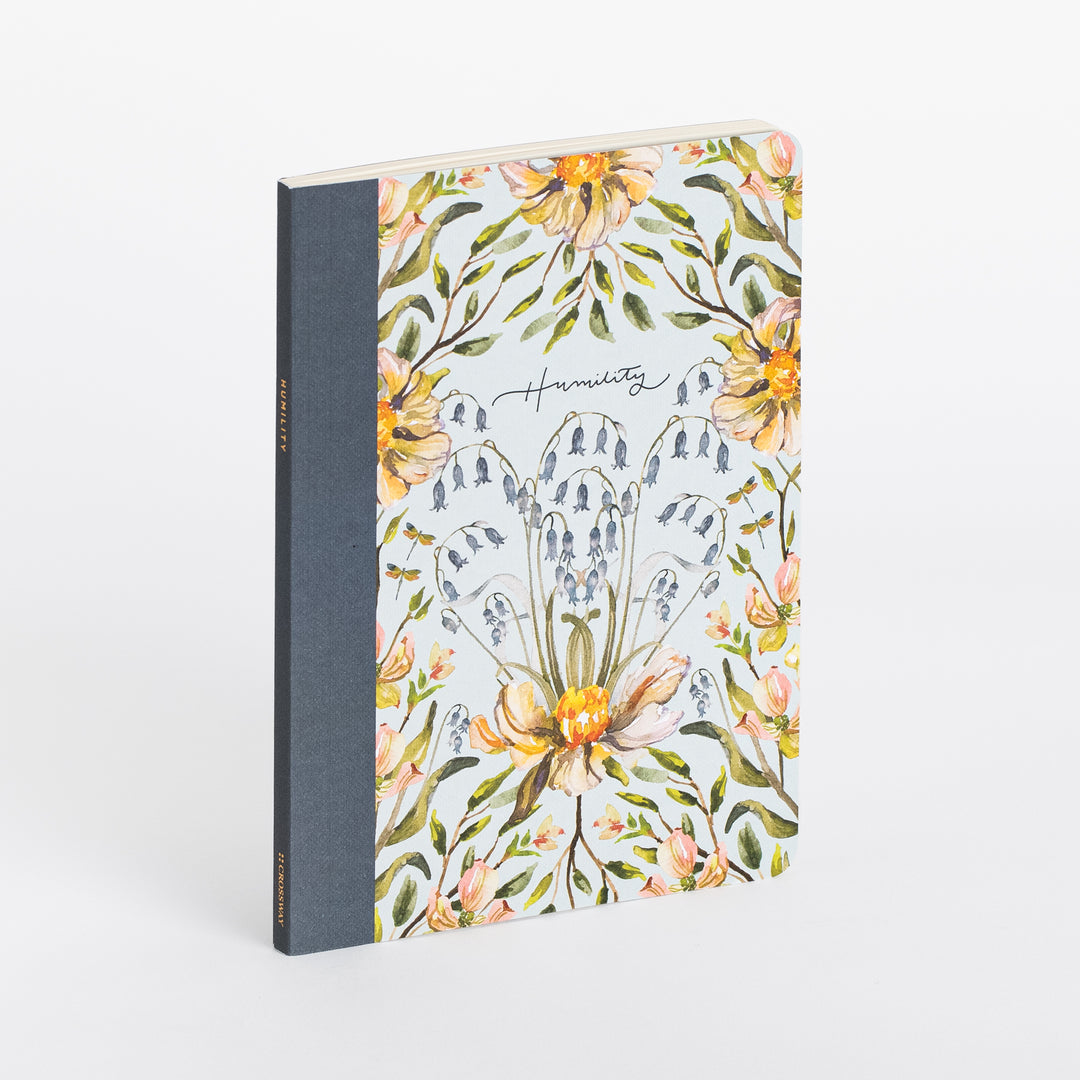 Softcover Humility scripture journal standing upright at an angle, showing a light blue cover with watercolor peach peonies, blue bellflowers, and green leaves surrounding a cursive 'Humility' title, and a slate-blue cloth spine reading HUMILITY / SCRIPTURE JOURNAL in gold foil.