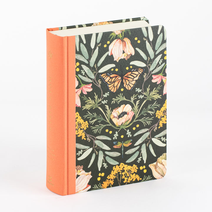 Hardcover Ruth Chou Simons Artist Series Bible standing at an angle, its front cover a dark charcoal ground with a symmetrical watercolor pattern of sage leaves, blush and cream flowers, yellow wildflower clusters, and a monarch butterfly at the center. The spine is burnt orange cloth with gold foil lettering, and the page edges are cream.