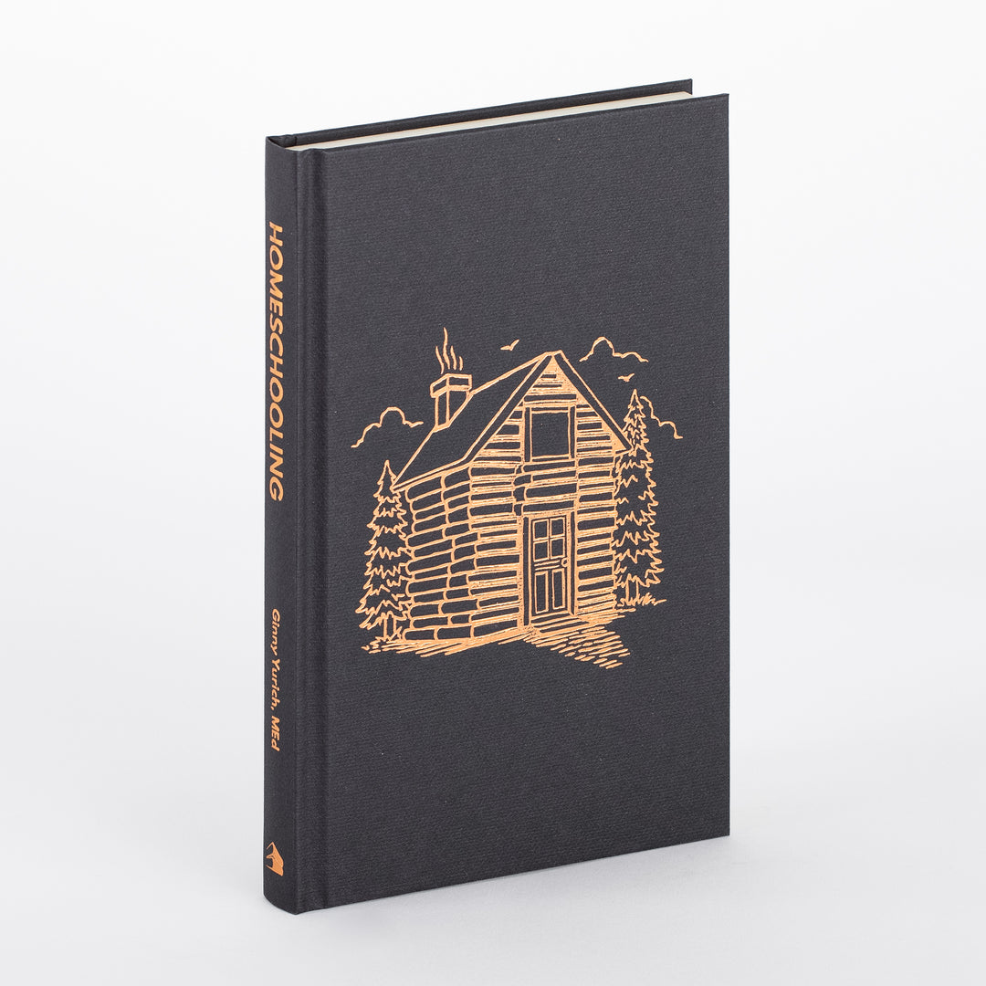 Homeschooling book with its dust jacket removed, showing the dark charcoal cloth hardcover case stamped in copper foil with the same log-cabin illustration and &