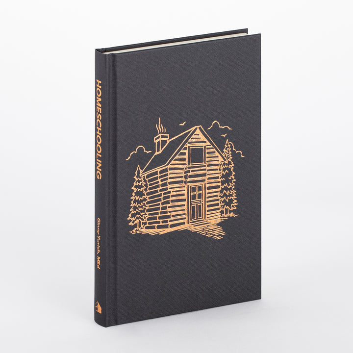 Homeschooling book with its dust jacket removed, showing the dark charcoal cloth hardcover case stamped in copper foil with the same log-cabin illustration and &