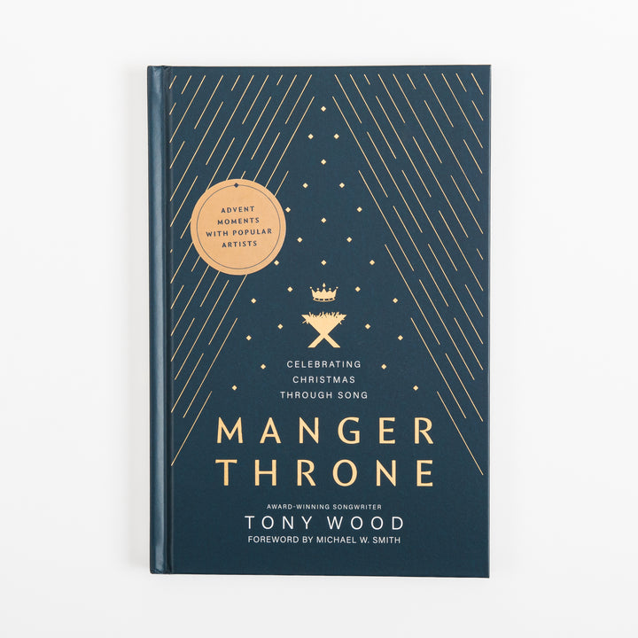 Straight-on front cover of the navy "Manger Throne" hardcover book with gold foil star lines, a gold crown-and-manger icon, a round "Advent Moments with Popular Artists" badge, and the title "Manger Throne: Celebrating Christmas through Song" by Tony Wood, foreword by Michael W. Smith.