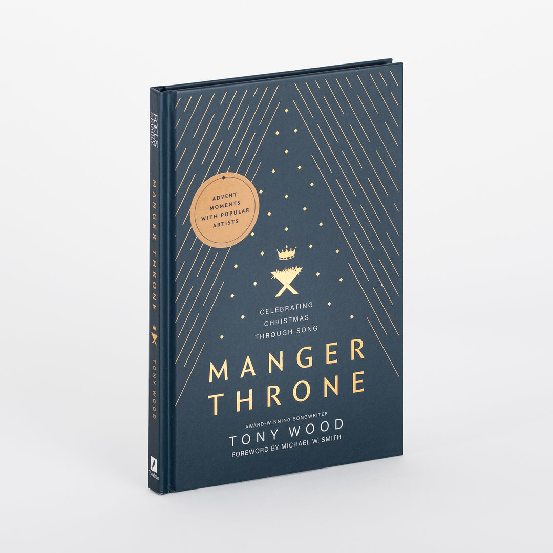 Angled view of the navy "Manger Throne" hardcover book, showing gold foil radiating lines, a small gold crown-over-manger icon, and a round tan badge reading "Advent Moments with Popular Artists," with title "Manger Throne: Celebrating Christmas through Song" by Tony Wood, foreword by Michael W. Smith.
