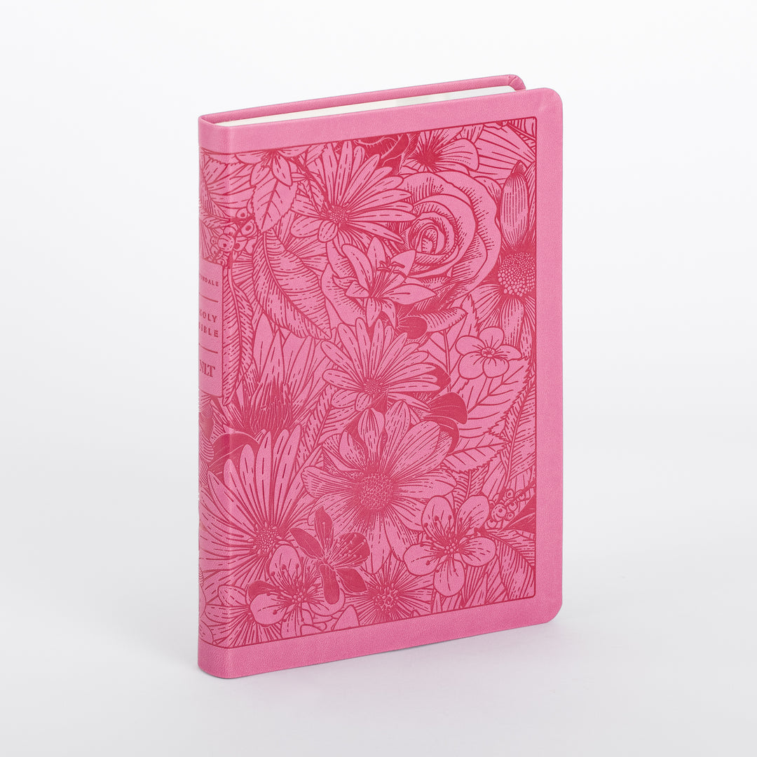 Pink leatherlike Bible cover shown at an angle, revealing both the front panel and spine. The front is densely engraved with a darker pink floral design of a large rose, daisies, and leaves inside a stitched border, while the spine shows a lighter pink title panel with partial lettering for the Holy Bible NLT.