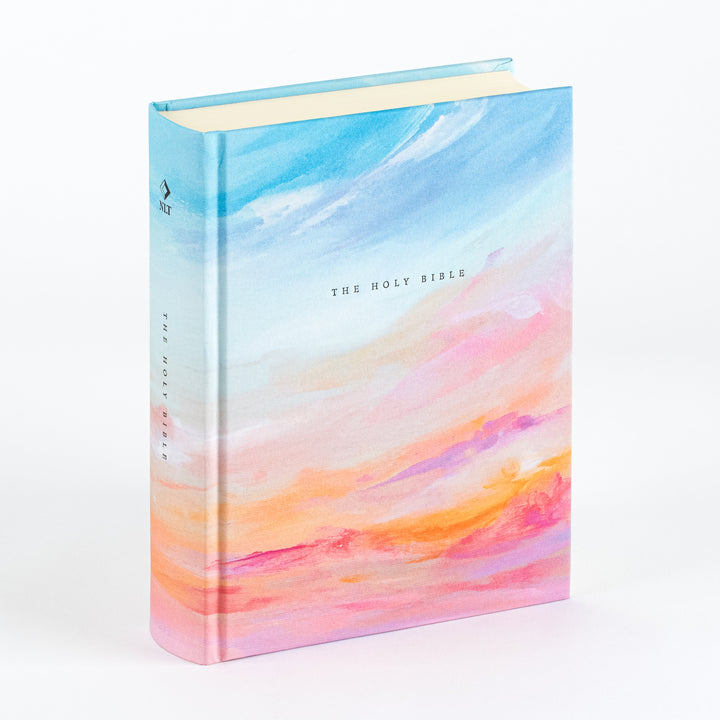 Angled view of the NLT Notetaking Bible in Rosemary Beach cloth-over-board cover, showing a hand-painted watercolor sky in soft blues fading into pink, orange, and purple sunset tones, with "THE HOLY BIBLE" printed in small serif type on the front cover and spine, an NLT diamond logo near the spine top, and cream-colored page edges.