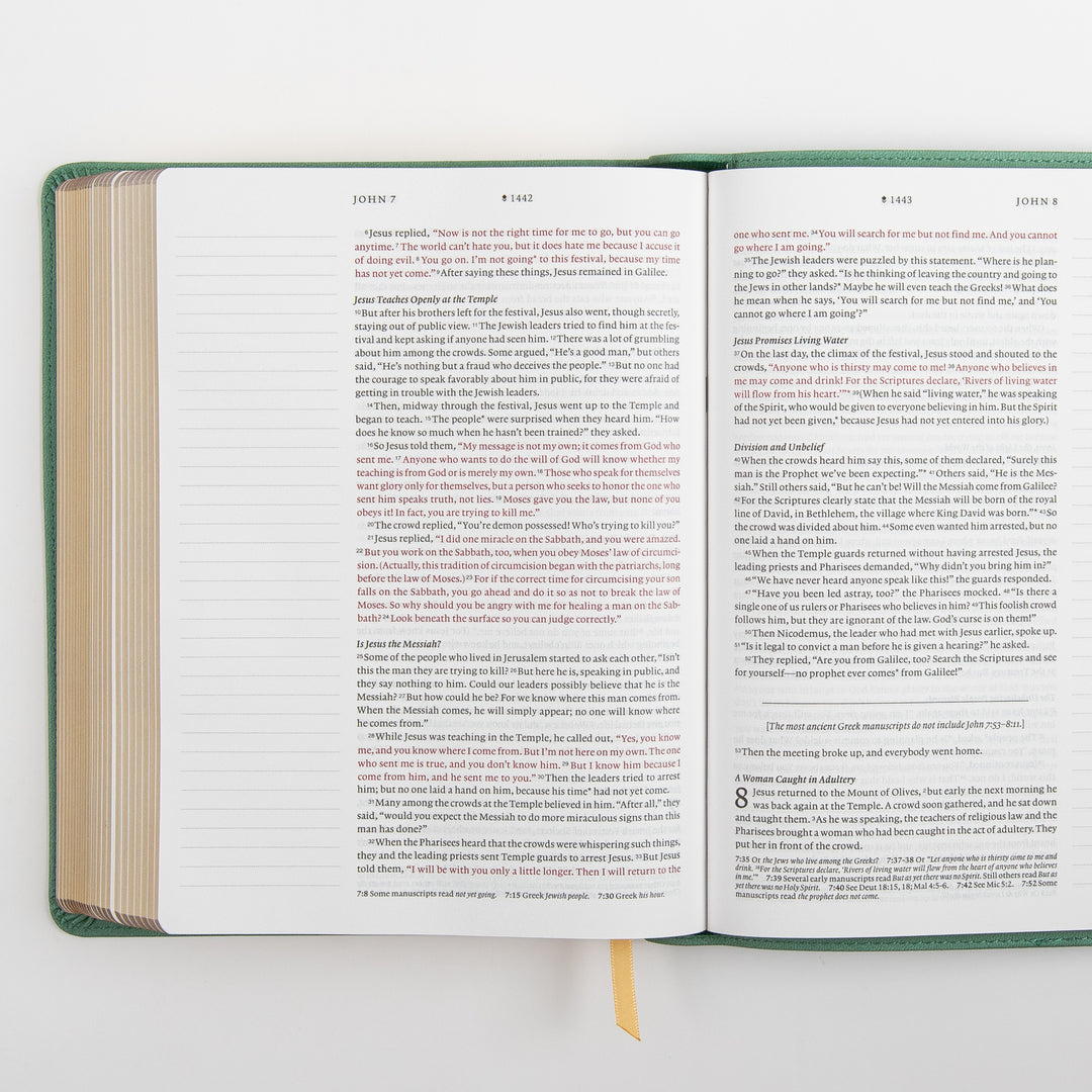 Angled close-up of the NLT Wide Margin Bible open to John 7-8, showing the account of the woman caught in adultery beginning at the bottom of the right page, with the words of Jesus printed in red and highlighted study-note callouts within the text.