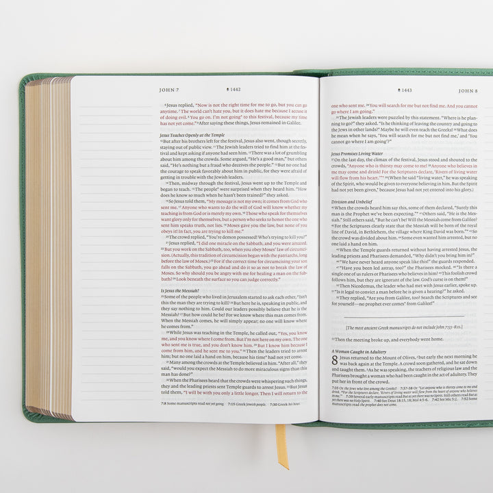 Angled close-up of the NLT Wide Margin Bible open to John 7-8, showing the account of the woman caught in adultery beginning at the bottom of the right page, with the words of Jesus printed in red and highlighted study-note callouts within the text.