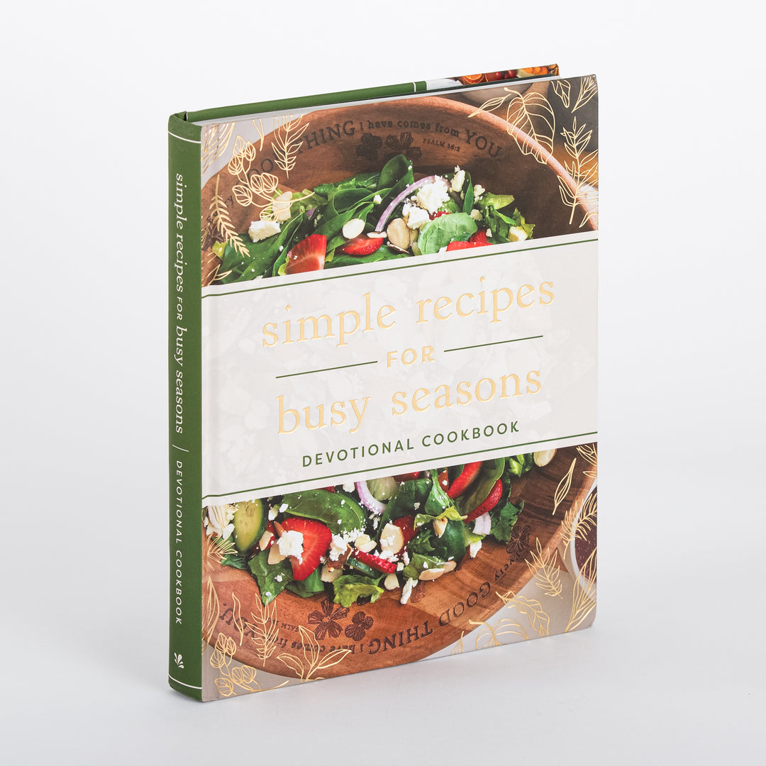 Angled three-quarter view of Simple Recipes for Busy Seasons Devotional Cookbook standing upright, showing the green spine reading simple recipes for busy seasons devotional cookbook beside the front cover's spinach salad photo and gold foil title.