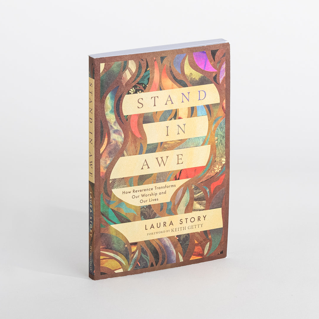 Angled view of the paperback cover of 'Stand in Awe: How Reverence Transforms Our Worship and Our Lives' by Laura Story, foreword by Keith Getty, against a white background. The cover has a colorful abstract stained-glass pattern of vines and leaves in teal, orange, purple, and gold, with the title on cream ribbon banners.