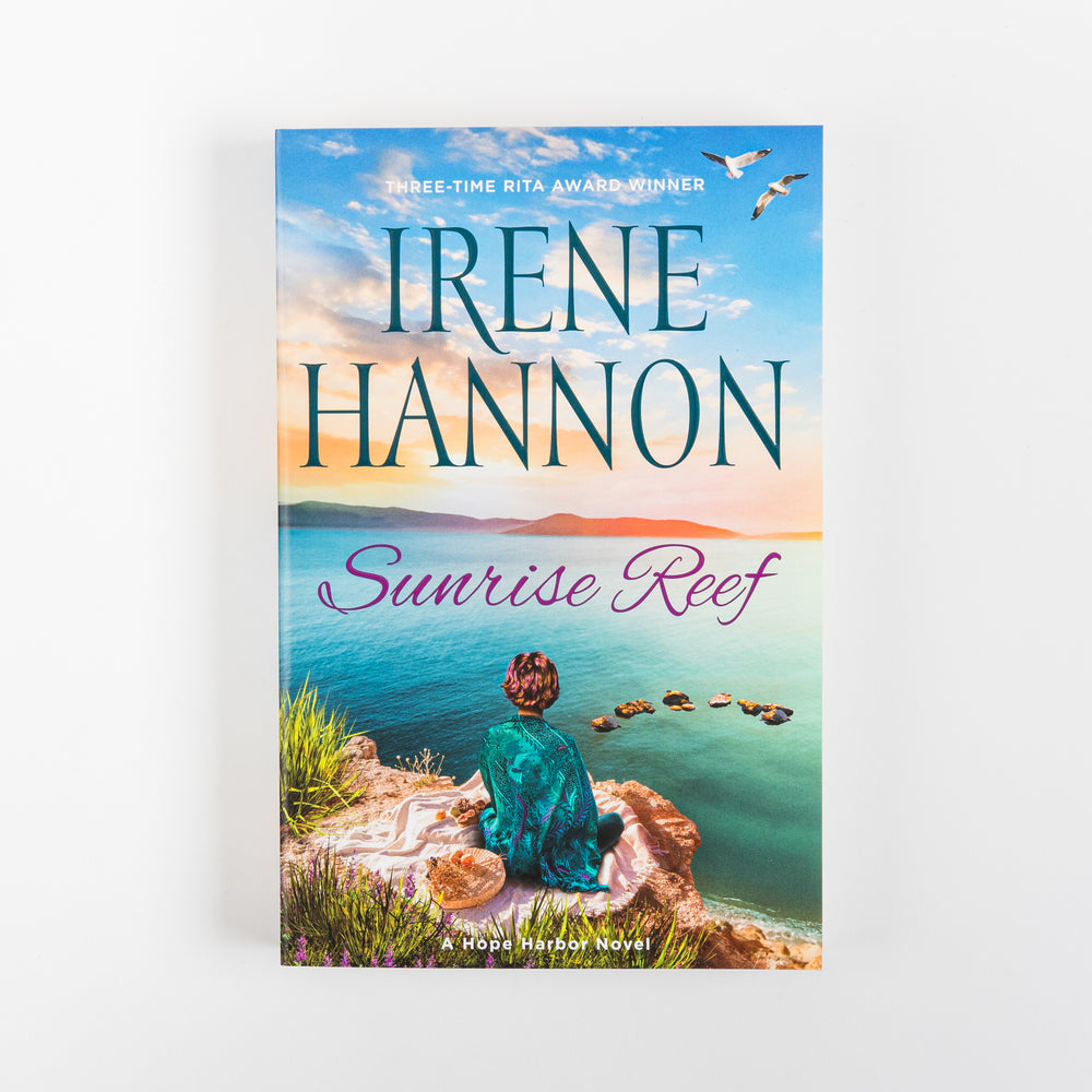 Front cover of the paperback novel Sunrise Reef by Irene Hannon, part of the Hope Harbor Novel series. A woman with red hair in a teal wrap sits facing away on a rocky ocean bluff at sunrise, with two seagulls flying overhead and a line of stacked stones leading into turquoise water. Title appears in purple script below the author's name.