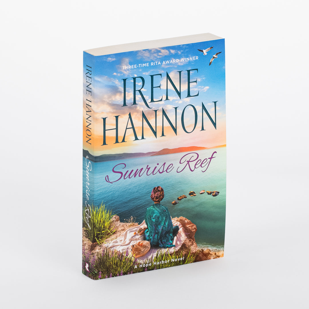 Angled 3D view of the paperback cover for Sunrise Reef by Irene Hannon, a Hope Harbor Novel. A woman in a teal wrap sits on a rocky coastal bluff overlooking a turquoise sea at sunrise, with two seagulls flying above and a trail of stacked stones leading into the water. Banner text reads Three-Time RITA Award Winner above the title in purple script.