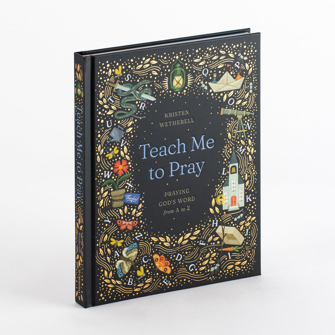 Hardcover book 'Teach Me to Pray: Praying God's Word from A to Z' by Kristen Wetherell, angled to show its dark navy cover and spine. Gold vines, dots, and hand-painted A-to-Z icons (lantern, church, snake, paper boat, telescope, potted flower, butterflies, carrot) frame the blue title and gold italic subtitle.
