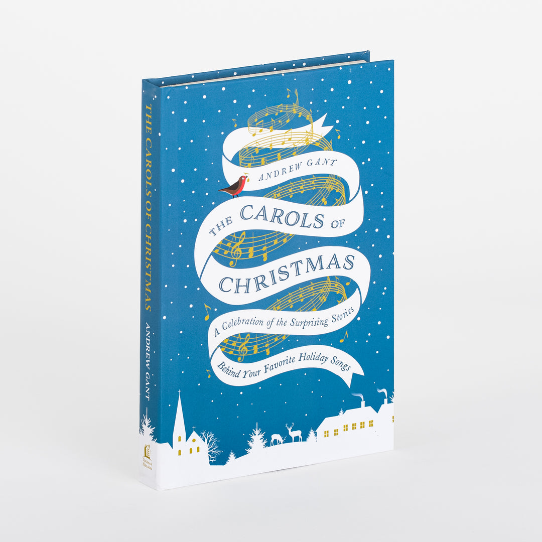 Three-quarter angled view of the hardcover book 'The Carols of Christmas' by Andrew Gant, showing its teal-blue cover and spine. A white ribbon banner threaded with gold musical staff lines and notes displays the title and subtitle, with a small red robin perched near the author's name. White silhouettes of a church, reindeer, and snow-covered cottages with lit windows run along the bottom edge.