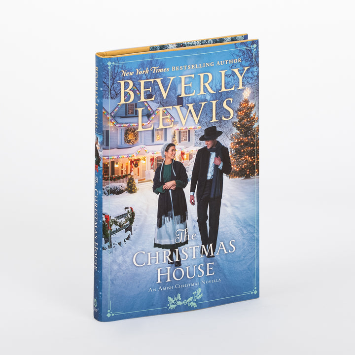 Hardcover of The Christmas House by Beverly Lewis, angled three-quarter view. The blue dust jacket shows an Amish woman and man walking arm in arm on a snowy path past a lit-up farmhouse and glowing Christmas tree at dusk.