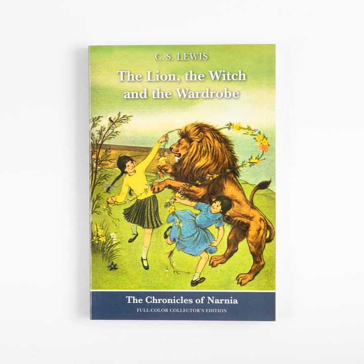 Front cover of the full-color paperback edition of The Lion, the Witch and the Wardrobe, showing two girls placing a flower wreath around the neck of Aslan the lion in a grassy field.