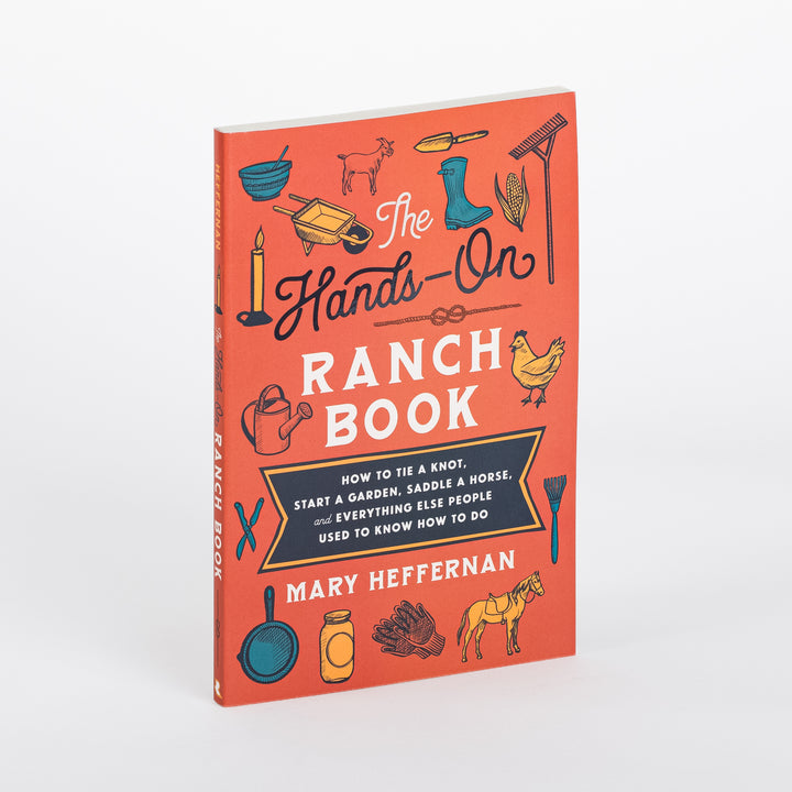 Angled view of The Hands-On Ranch Book by Mary Heffernan: an orange paperback cover with hand-drawn illustrations of a goat, chicken, horse, wheelbarrow, corn, boots, rake, candle, watering can, skillet, mason jar, and gloves around the title and the subtitle &