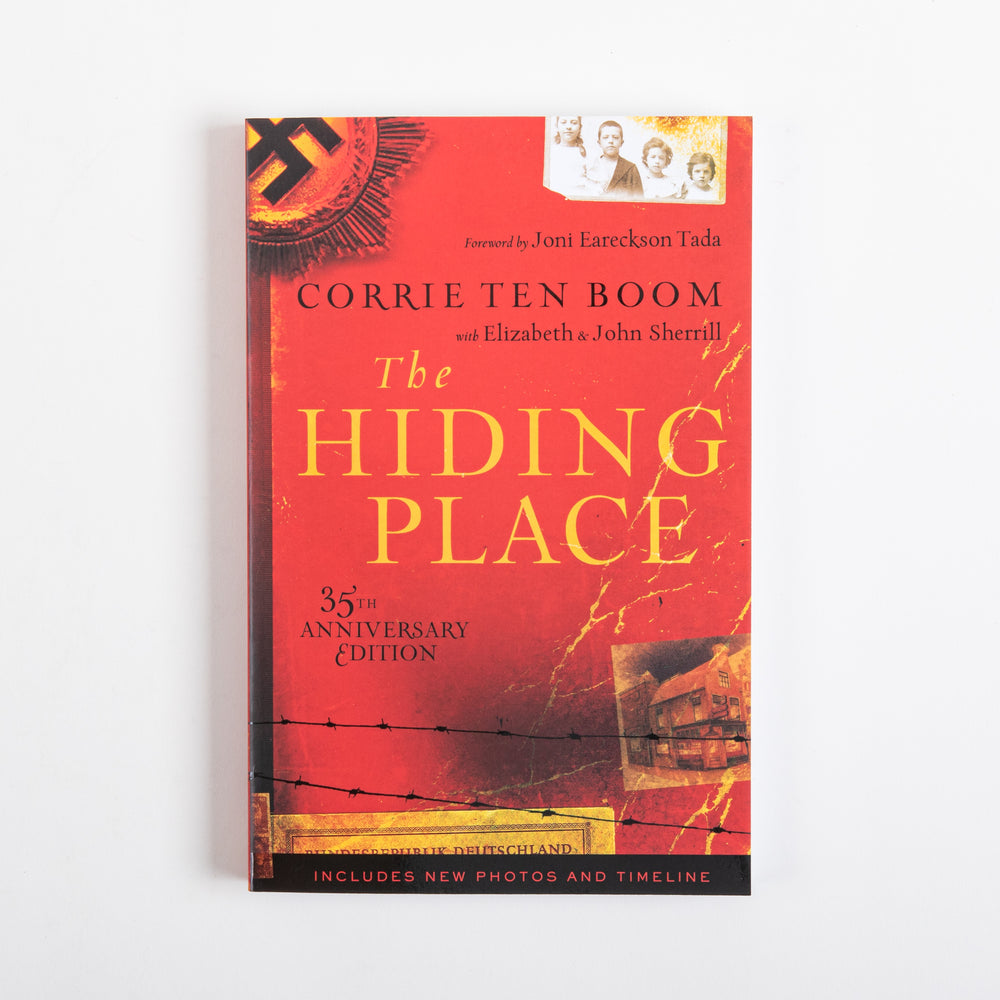 Front cover of the paperback The Hiding Place, 35th Anniversary Edition by Corrie ten Boom with Elizabeth and John Sherrill, foreword by Joni Eareckson Tada. Red background with a black Nazi emblem graphic top left, a sepia family photo top right, gold title lettering, barbed wire illustration, a sepia building photo, and a black bottom band reading Includes New Photos and Timeline.