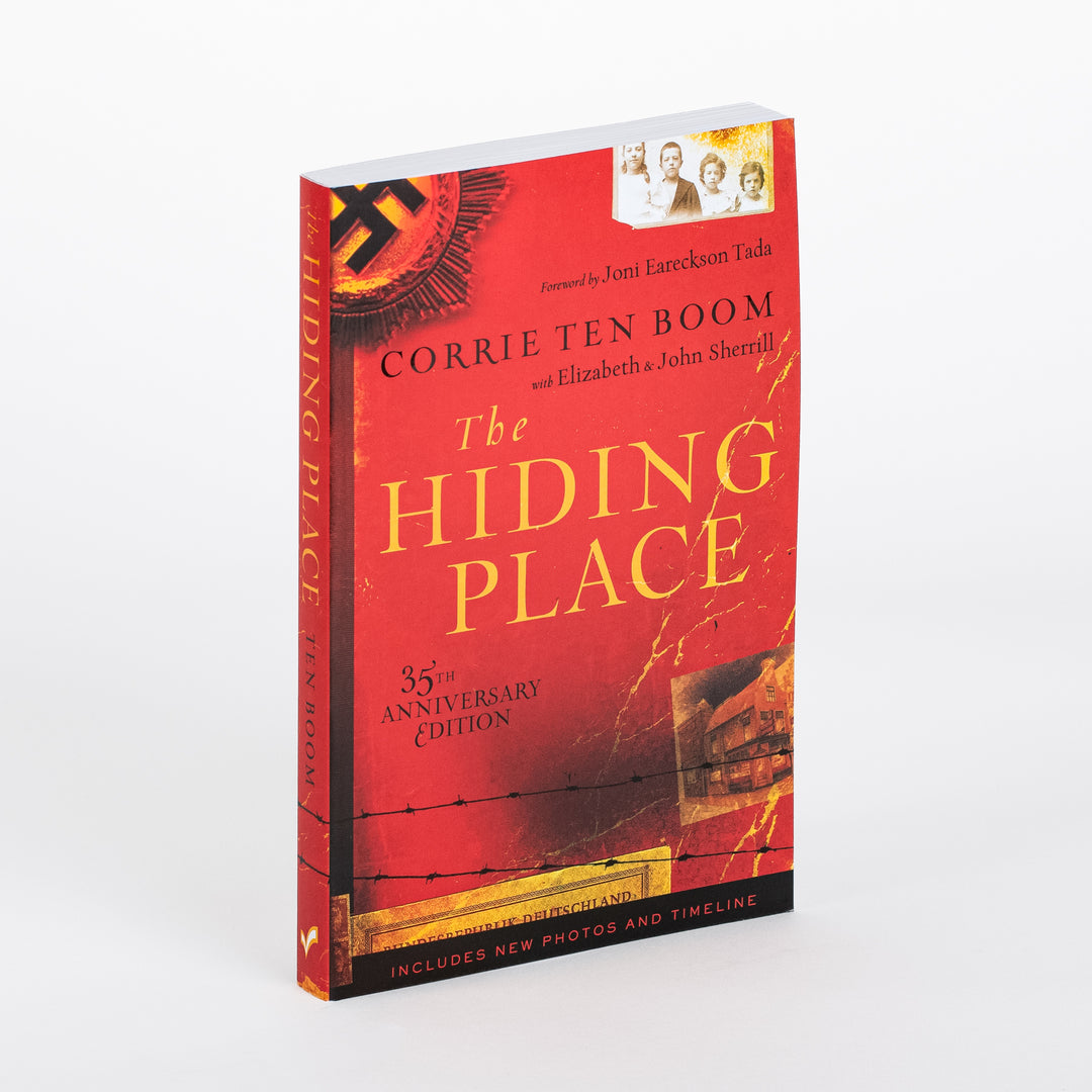 Angled three-quarter view of the paperback book The Hiding Place, 35th Anniversary Edition by Corrie ten Boom with Elizabeth and John Sherrill, foreword by Joni Eareckson Tada. Red cover shows a black Nazi emblem graphic, a sepia family photo, gold title text, barbed wire art, and a bottom black band reading Includes New Photos and Timeline; the book's spine is visible at left.