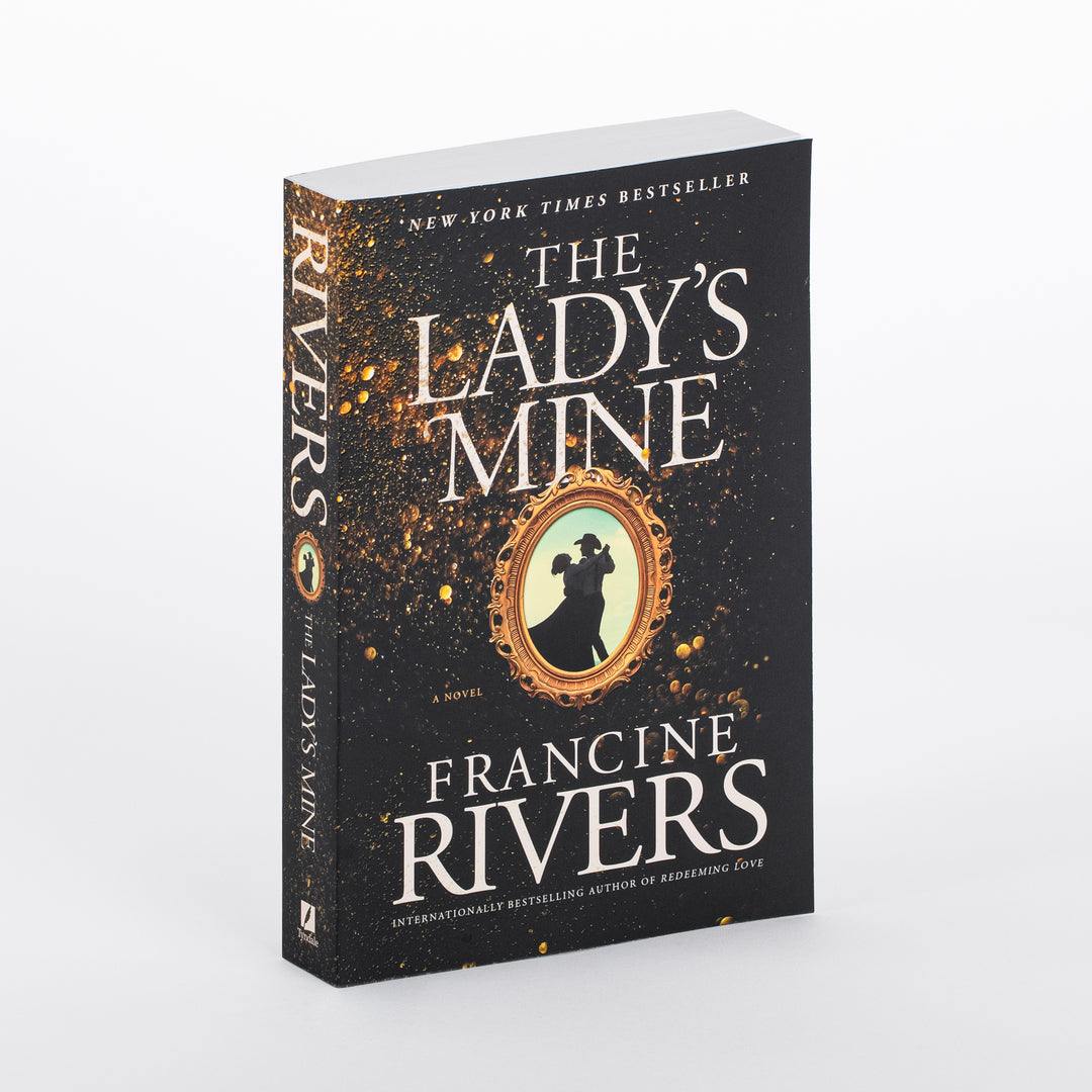 Angled three-quarter view of the paperback novel The Lady's Mine by Francine Rivers, showing both front cover and spine. The black cover is speckled with gold paint flecks radiating from a center gold oval frame holding the black silhouette of a couple dancing, the man in a wide-brimmed hat. White serif text reads 'New York Times Bestseller' at top, then the title 'The Lady's Mine,' and 'Francine Rivers' in large letters near the bottom. The spine repeats 'Rivers' and the title vertically.