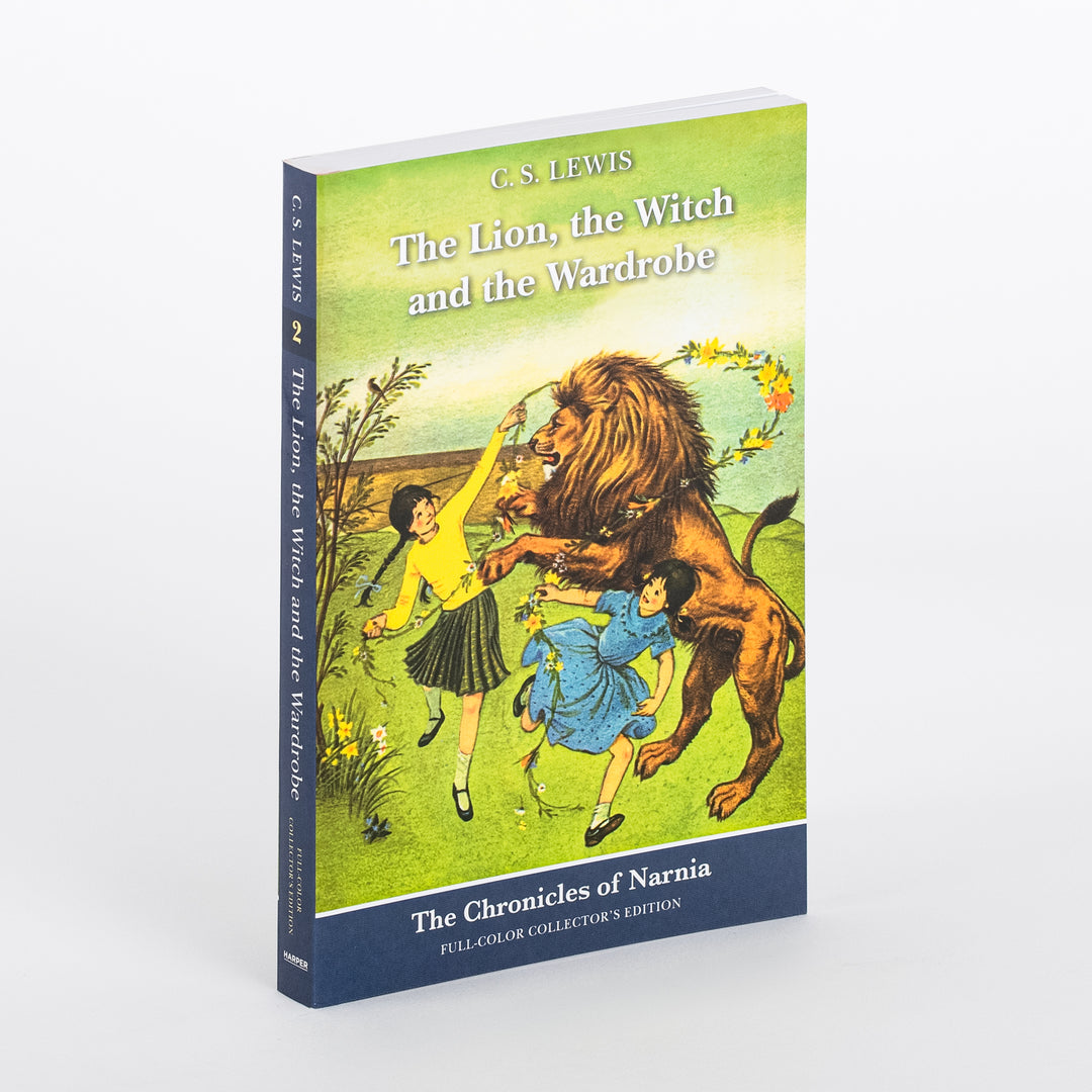 Angled three-quarter view of 'The Lion, the Witch and the Wardrobe: Full-Color Collector's Edition' paperback by C.S. Lewis, showing the front cover illustration of two girls draping a flower garland on a lion in a green field beneath a pale sky, with the navy spine reading the title and 'The Chronicles of Narnia' visible at left.