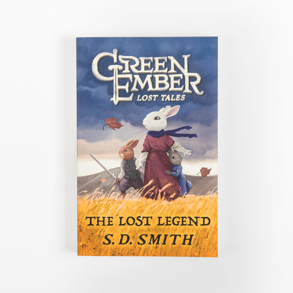 Front cover of 'The Lost Legend (Green Ember Lost Tales 2)' by S. D. Smith, shown flat. A white rabbit in a maroon cloak and blue scarf stands over two young rabbits, one armed with a sword, in a golden wheat field beneath a dark blue stormy sky with leaves blowing in the wind.