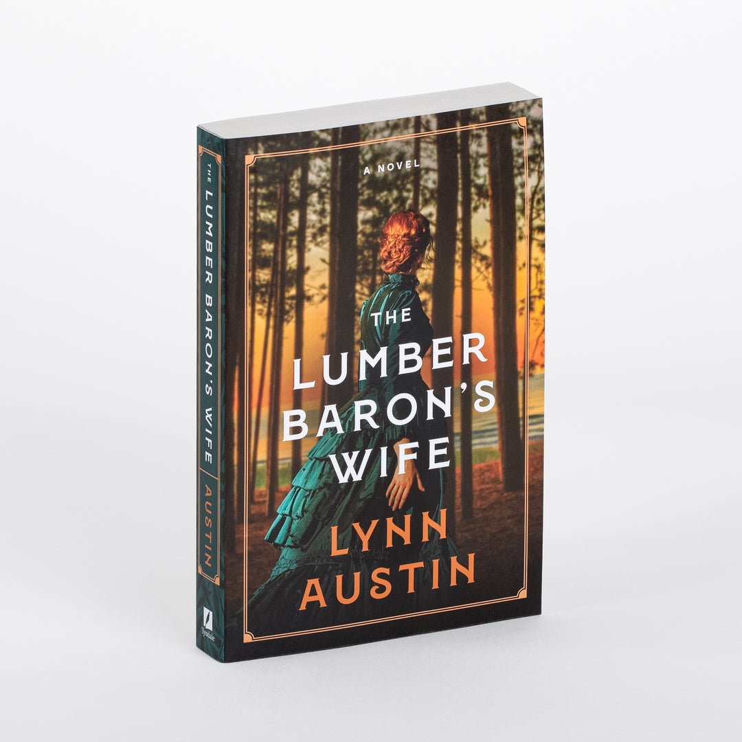 Paperback book shown at a three-quarter angle, revealing both the front cover and spine of 'The Lumber Baron's Wife' by Lynn Austin. The cover art shows a red-haired woman in a teal Victorian dress with a ruffled bustle, seen from behind, walking through dark pine tree trunks silhouetted against a glowing orange sunset over water. 'A Novel,' the bold white title, and the orange author name sit inside a thin gold border; the dark green spine repeats the title and author vertically.