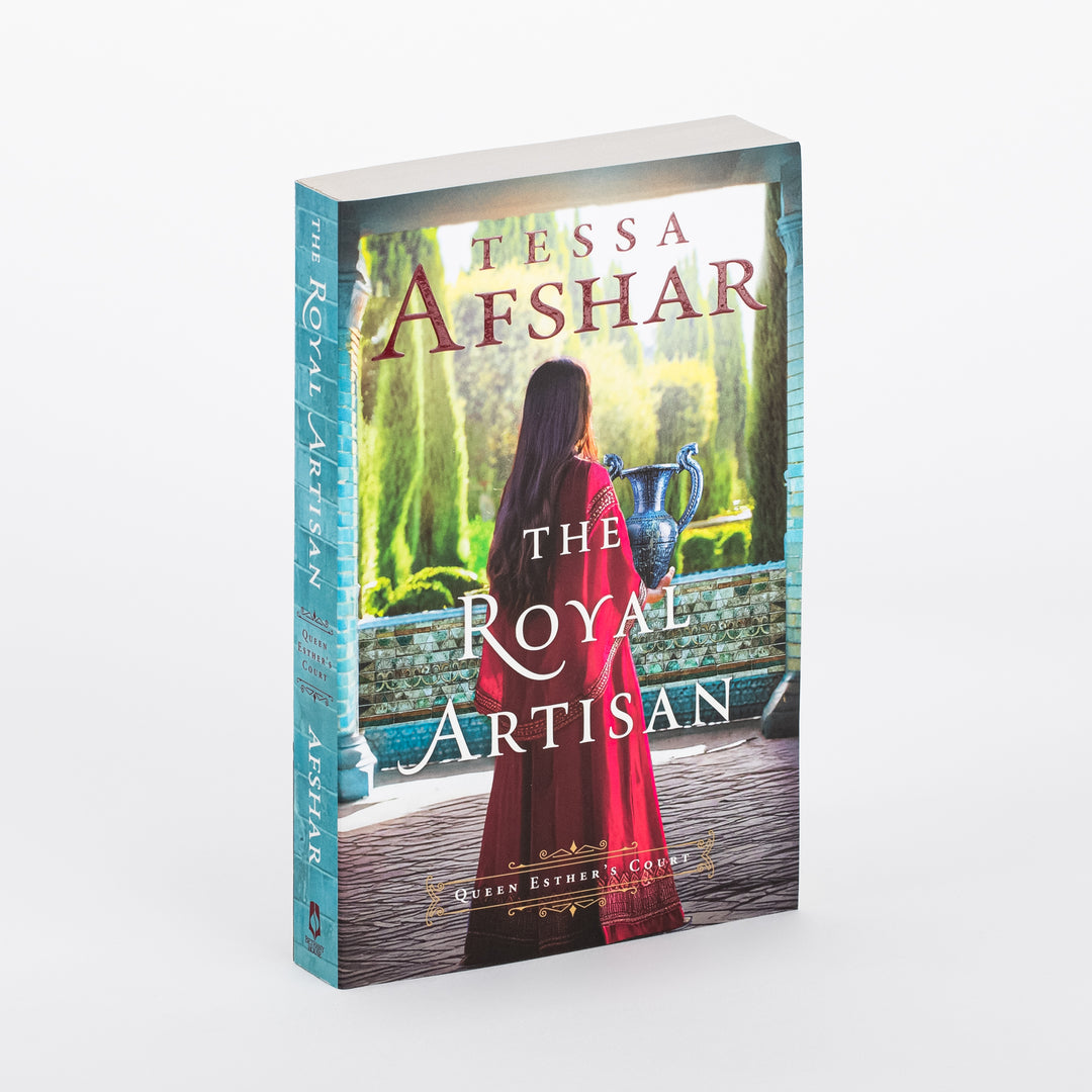 Paperback cover of The Royal Artisan by Tessa Afshar, shown at an angle. A woman in a flowing red robe, seen from behind, holds a blue and silver ceramic vase while standing at a turquoise-tiled archway overlooking a green garden. Queen Esther's Court series label near the bottom.