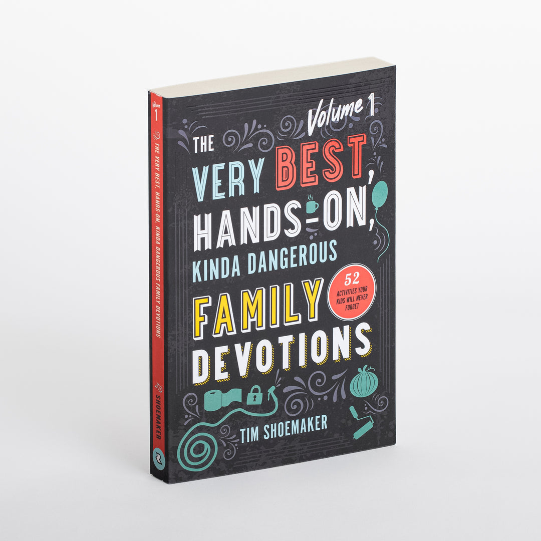 Paperback book angled in 3/4 view on white background: black cover reading 'The Very Best, Hands-On, Kinda Dangerous Family Devotions, Volume 1' by Tim Shoemaker, with a red circular badge noting '52 activities your kids will never forget' and teal doodle icons of a balloon, toilet paper roll, lock, and pumpkin.