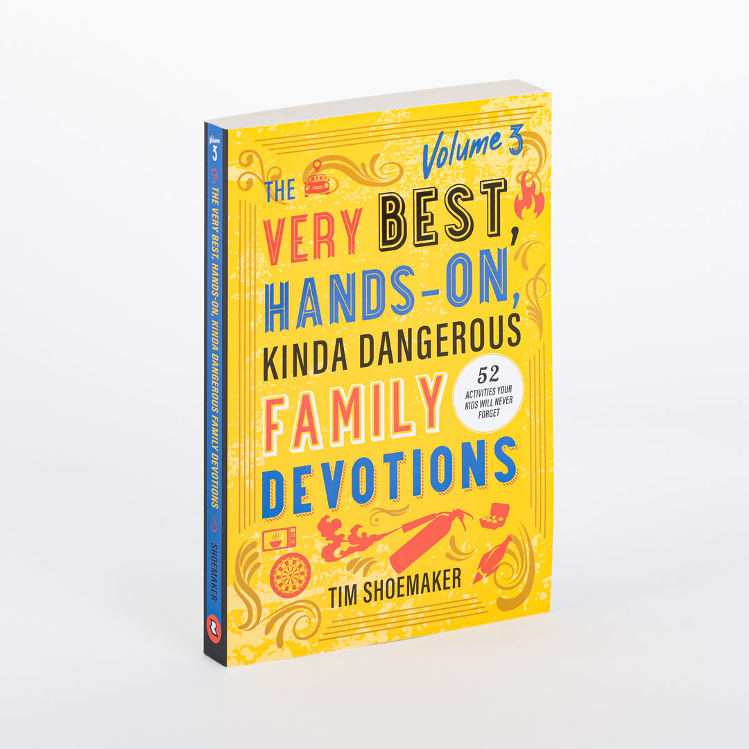 Paperback book cover at an angle showing the spine and front of 'The Very Best, Hands-On, Kinda Dangerous Family Devotions, Volume 3' by Tim Shoemaker. Bright yellow cover with red, blue, and black hand-lettered title text, a white '52 Activities Your Kids Will Never Forget' badge, and illustrated icons including a school bus, flame, dart board, and spray can.