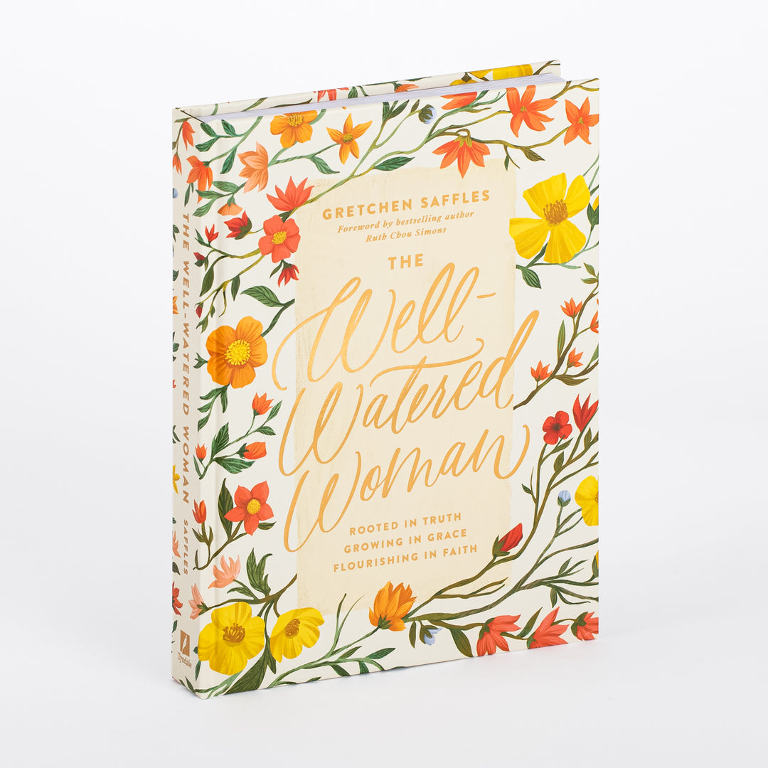 Hardcover book The Well-Watered Woman by Gretchen Saffles shown at an angle, revealing its spine and front cover illustrated with yellow, orange, and red wildflowers on a cream background, gold script title, and subtitle Rooted in Truth, Growing in Grace, Flourishing in Faith.