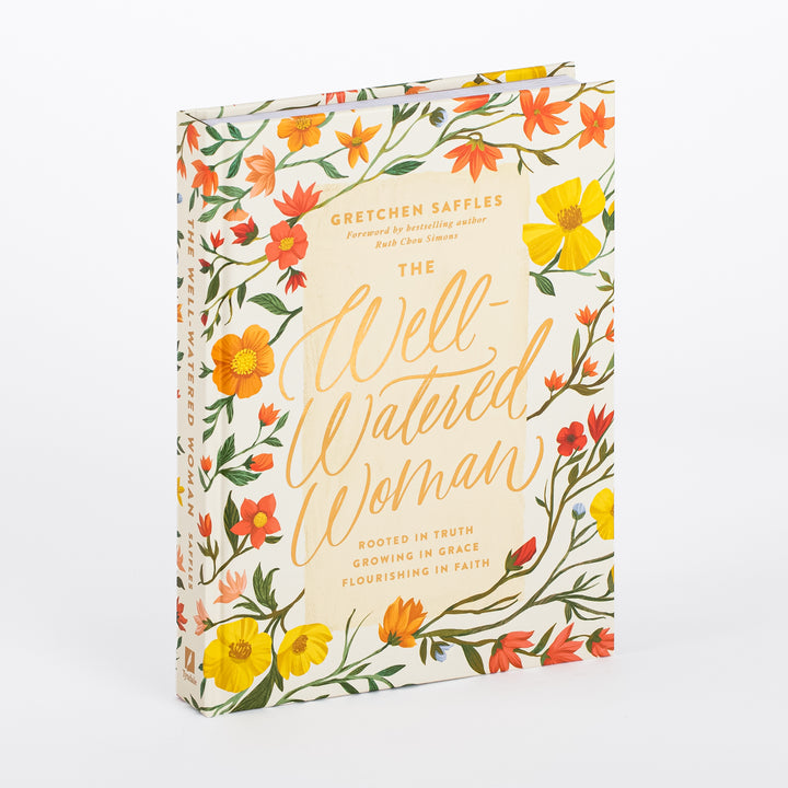 Hardcover book The Well-Watered Woman by Gretchen Saffles shown at an angle, revealing its spine and front cover illustrated with yellow, orange, and red wildflowers on a cream background, gold script title, and subtitle Rooted in Truth, Growing in Grace, Flourishing in Faith.