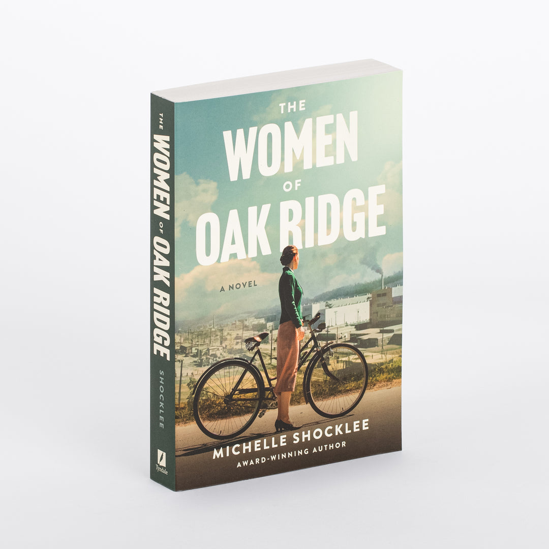 Angled three-quarter view of 'The Women of Oak Ridge' by Michelle Shocklee paperback, showing the spine and front cover: a woman in a green cardigan stands beside a bicycle on a road, looking toward a hazy 1940s industrial town with a smoking factory chimney under a teal sky.