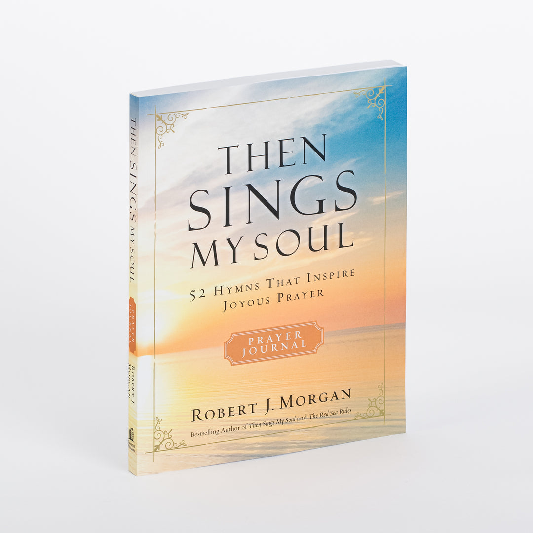 Angled three-quarter view of the Then Sings My Soul Prayer Journal: 52 Hymns That Inspire Joyous Prayer softcover book by Robert J. Morgan, showing its thickness and spine against a sunset-over-ocean cover design with gold corner ornaments and an orange Prayer Journal badge.