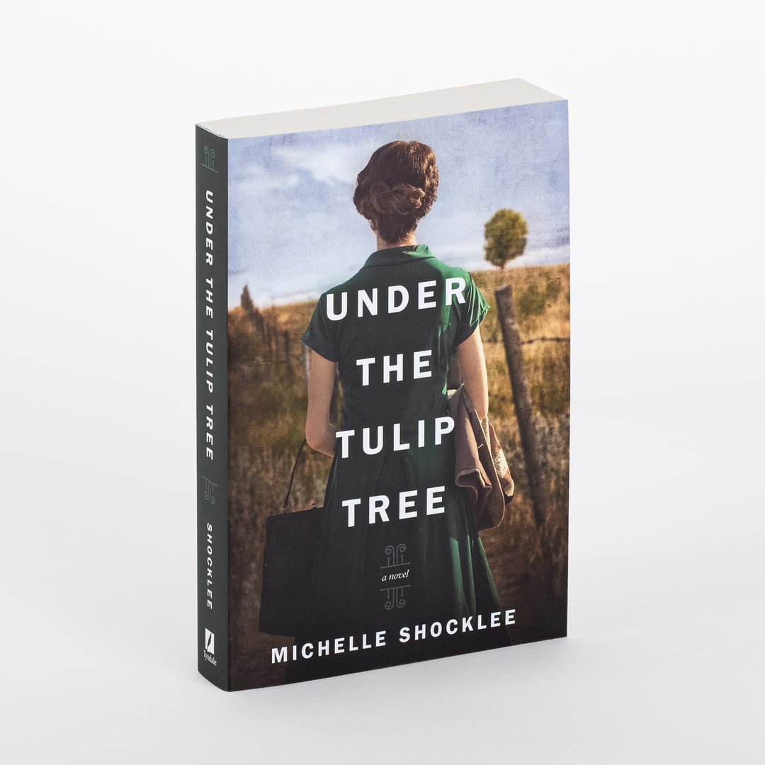 Angled view of 'Under the Tulip Tree' by Michelle Shocklee showing spine and front cover: a woman in a green vintage dress with an updo stands with her back to viewer beside a wooden fence in a golden field, a lone round-topped tree in the distance.