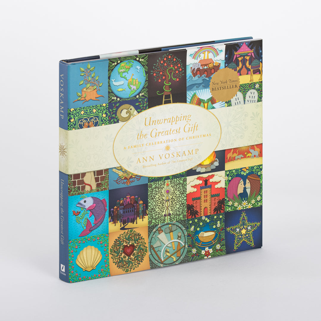 Three-quarter angle view of the hardcover Unwrapping the Greatest Gift: A Family Celebration of Christmas by Ann Voskamp, standing upright with its spine reading VOSKAMP and the same colorful folk-art icon quilt cover design wrapping around the front, including the New York Times Bestseller seal.