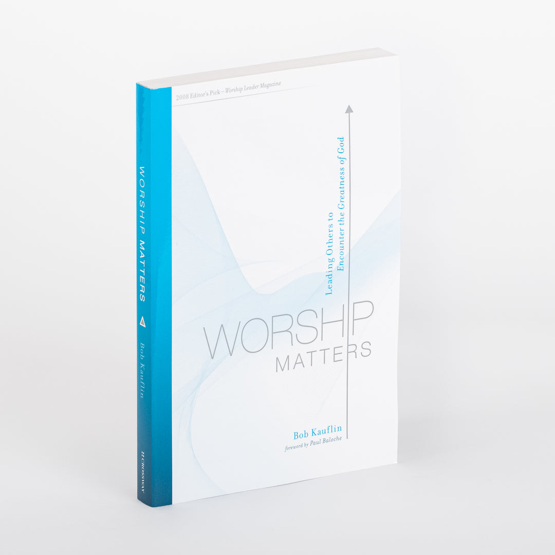 Angled three-quarter view of the paperback 'Worship Matters: Leading Others to Encounter the Greatness of God' by Bob Kauflin, foreword by Paul Baloche. White cover with a pale blue abstract wave graphic and thin vertical arrow beside the vertical subtitle text; teal-to-blue gradient spine visible at left, on a white background.